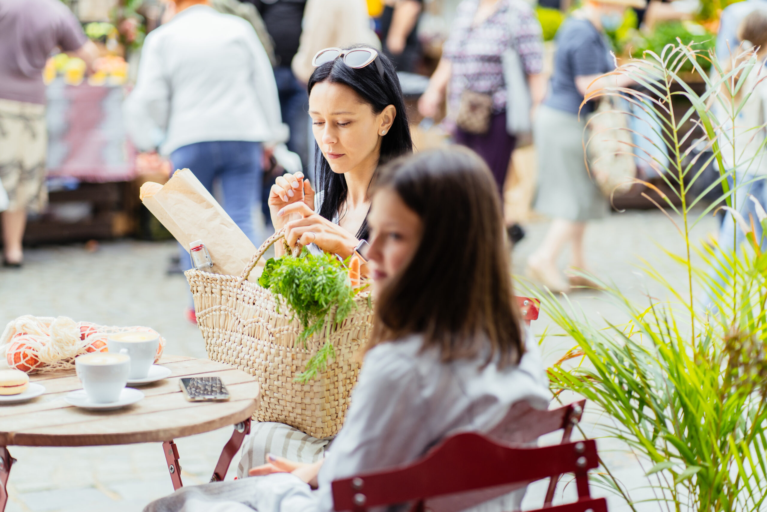 Brunette slim sensual mother and her teenager daughter relaxing after shopping at farmer market, sitting at outdoor cafe with wicker bag full of fruits, green, vegetables. Shopping, eco food concept.